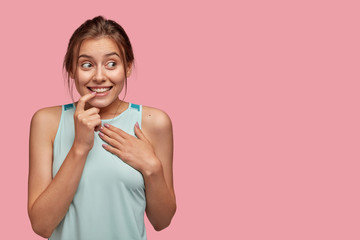 Isolated shot of positive European woman bites finger, keeps hand on chest, smiles positively, looks aside, wears casual clothes, hears intriguing information, isolated over pink wall with free space
