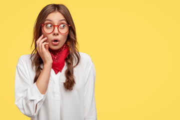 Oops, it cant be so. Stupefied European woman in spectacles, red bandana and white shirt, looks...