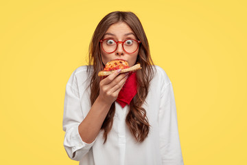 Surprised European fashionable woman has slice of pizza, looks at camera, dressed in oversized...