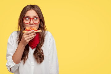 Satisfied Caucasian model eats delicious pizza indoor, has lunch, wears optical glasses, white shirt and red bandana, stands against yellow background with free space for your slogan or text