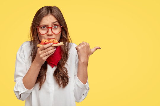 Horizontal shot of surprised female eats tasty slice of pizza, dressed in fashionable clothes, indicates with thumb, invites you to pizzeria, isolated over yellow studio wall. People and nutrition