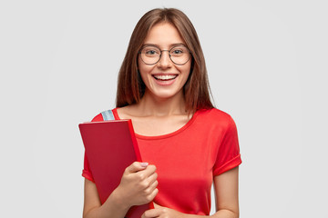 Positive Caucasian girl with charming smile, wears red t shirt, holds textbook, models against...