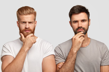 Photo of serious two men hold chins, dressed in casual t shirts, model against white background,...