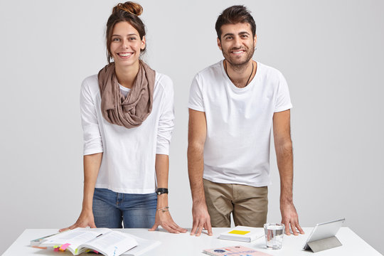 Photo of glad young female and male students lean at table, stand at desktop, learn information, use books and tablet, isolated over white background, solve issues, have brainstroming session