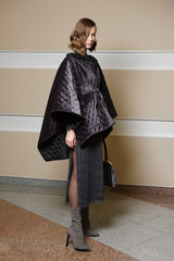 Full length portrait of beautiful girl in knitted dress and poncho coat. Gorgeous lady in warm outfit with hand bag posing near the wall