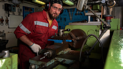 Worker in red uniform operating in manual lathe in metal big workshop.