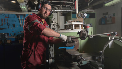 Worker in red uniform operating in manual lathe in metal big workshop.