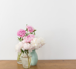 Pink and white peonies in glass and green vases on wooden table against wall (selective focus)
