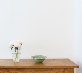 Pale pink peonies in glass vase with green bowl on wooden sidetable against white wall (selective focus)