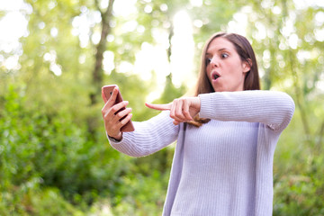 Naklejka premium Young caucasian european brunette girl pointing her cellphone very surprised in the forest or nature