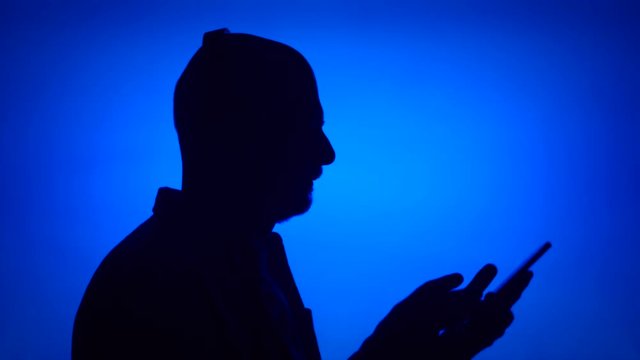 Silhouette of senior man listen to music on cellphone. Male's face in profile put on big black wireless headphones on blue background. Black contour shadow of grandfather's half-face singing