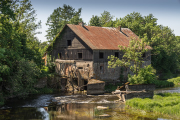 Fototapeta na wymiar Ancient abandoned water mill surrounded by beautiful nature. House built of stone and wood, exterior walls and dilapidated bridge on river is reflection of trees and hous ruins