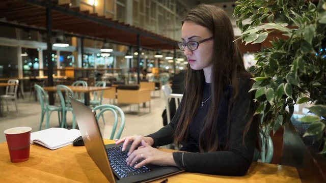 Young girl in spectacle glasses working with laptop and typing at a wooden table in an empty airport terminal cafe