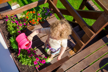 Top view of a young gardener girl watering pink flowers in flowerpot with children`s watering can on a brown terrace outdoors on sunny day. © FotoHelin