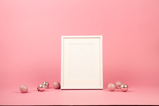 Mock up with white picture frame on trendy pastel light pink background with christmas ornaments. Greeting card and pink Christmas baubles