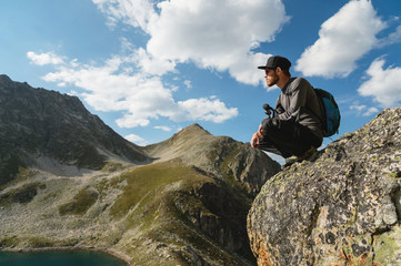 A bearded hipster tourist in sunglasses with a backpack sits on the edge of a cliff high in the mountains near the mountain lake