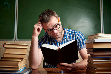 A disheveled tired unshaven young man with glasses holds his head and reads a book at the table...