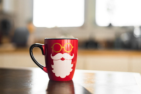 Cute Festive Holiday Photo Of Bright Red Seasonal Santa Christmas Coffee Mug with Hipster Beard and Hot Steam Coming Off Cup in Light White Modern Kitchen with Sunshine Such a Warm Cozy Morning