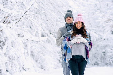 Fototapeta na wymiar Beautiful pregnant woman with her husband dressed grey knitted sweaters, hat, scarf and jeans, holding pink cups with hot tea and walking in a snowy winter park on background.