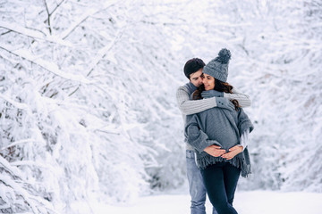 Fototapeta na wymiar Husband in a grey sweater and jeans tenderly hugging his beautiful pregnant wife in grey knitted sweaters, hat, scarf and jeans in a snowy winter park. Pregnancy concept.