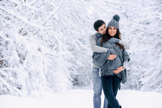 Husband in a grey sweater and jeans tenderly hugging his beautiful pregnant wife in grey knitted sweaters, hat, scarf and jeans in a snowy winter park. Pregnancy concept.