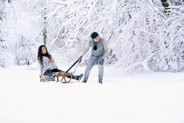 Young man rides his beautiful pregnant wife in wooden sledding in the snow in the park. Family love and care concept.