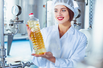 A young girl in white overalls is holding a bottle of sunflower oil in her hands against the background of the factory equipment of the food industry