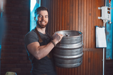 A young brewer in an apron holds a barrel with beer in the hands of a brewery