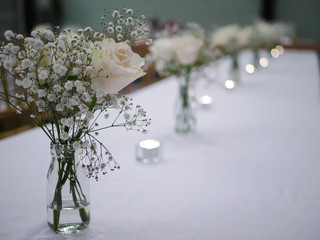 White rose and white flower with green stem in glass bottle jar vase on white table cloth with tea light candles