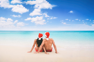 Christmas beach vacation holidays santa hat couple relaxing from behind sitting on white sand blue sky background for text advert for holiday season. © Maridav