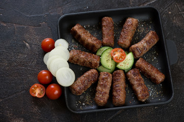 Cast-iron serving pan with grilled cevapi or cevapcici sausages, elevated view on a dark brown stone background