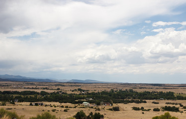 Landscape in the steppe, surrounded by mountains on a summer day. Houses stand at the foot of the mountains, dry grass on the ground, trees grow.