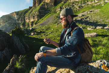 Naklejka na ściany i meble Close-up of a bearded man in jeans clothes in sunglasses and a cap with a backpack sitting at the foot of the epic rocks with a compass in his hands. Navigation travel tourism and orientation
