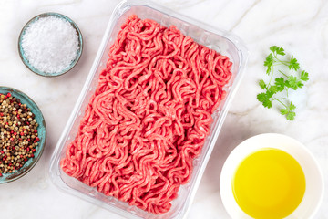 A photo of minced meat in a plastic tray, shot from the top on a white marble kitchen surface with salt, pepper, olive oil, and parsley leaves and copy space