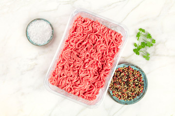 A photo of minced meat in a plastic tray, shot from above on a white marble background with salt,...