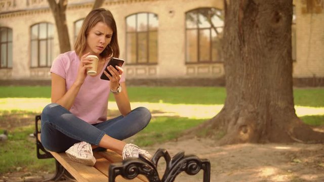 caucasian student sitting on the bench near university building. girl with blond hair sharing photo in social media drinking coffee