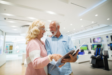 Happy senior couple looking at catalog while standing in tech store. Woman pointing at product.