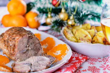 Baked meat in net on a white plate next to tangerine circles and a plate of baked potatoes on a red festive napkin on a New Year holiday table