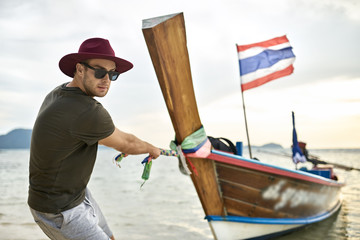 Man with stubble is pulling wooden boat by rope on shallow water