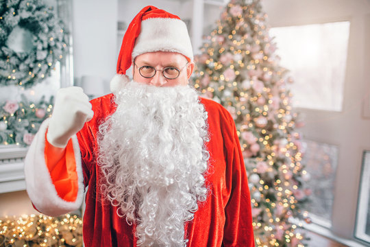 Picture of Santa Claus stands and looks on camera. He shows fist. Man threatens. He is in festive room.