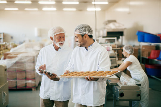 Controller talking to worker while using tablet. Worker carrying plate with cookies. Protective clothes on. Food factory interior.