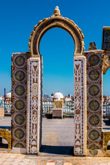 A Tunisian rooftop overlooking Tunis with a bright blue/aqua sky, archway in the foreground and mosque in the background.