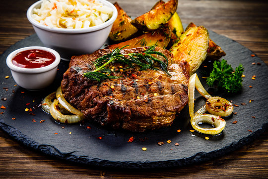 Grilled steak with baked potatoes and vegetables served on black stone plate on wooden table