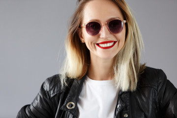 Beautiful young woman blonde in leather jacket and sunglasses smiles and poses in the studio on the grey background. Place for text