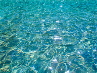 Blue transparent water surface with bright sun glare and sunny shining reflections on bottom. Top view of turquoise ripple texture with sunlight refracting through liquid layer.