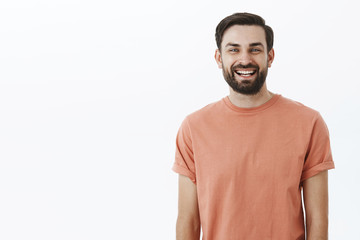 Fototapeta premium Waist-up shot of bright and cheerful emotive friendly bearded 30s man with broad smile gazing satisfied and upbeat at camera as posing in pink t-shirt over gray background, amused and enthusiastic