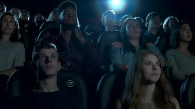 Wide shot of young movie theatre audience sitting, watching a movie. Slow dolly left to right.