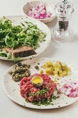 Beef tartare with quail egg in shell, cutting pickled cucumbers, capers, red onion, chives and arugula salad served in spotted ceramic plates with pepper mill on white marble table.