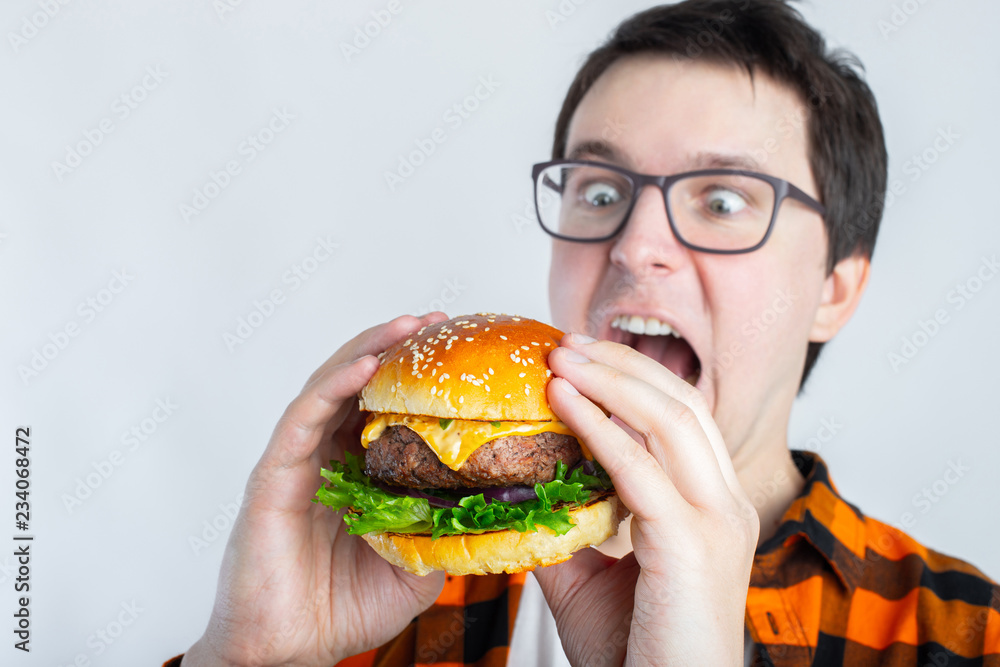 Wall mural A young guy with glasses holding a fresh Burger. A very hungry student eats fast food. Hot helpful food. The concept of gluttony and unhealthy diet. With copy space for text