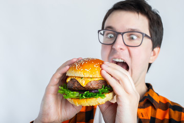A young guy with glasses holding a fresh Burger. A very hungry student eats fast food. Hot helpful...